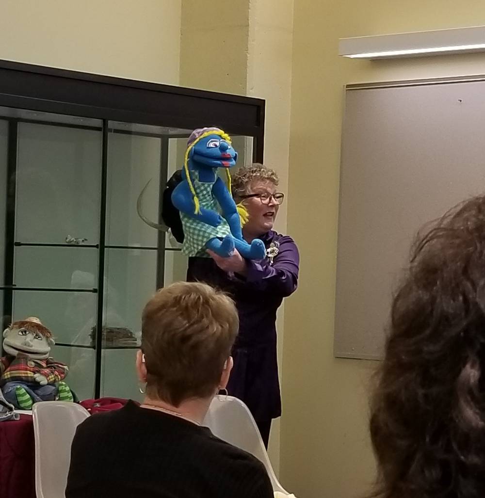 A woman holds up a blue puppet with yellow hair